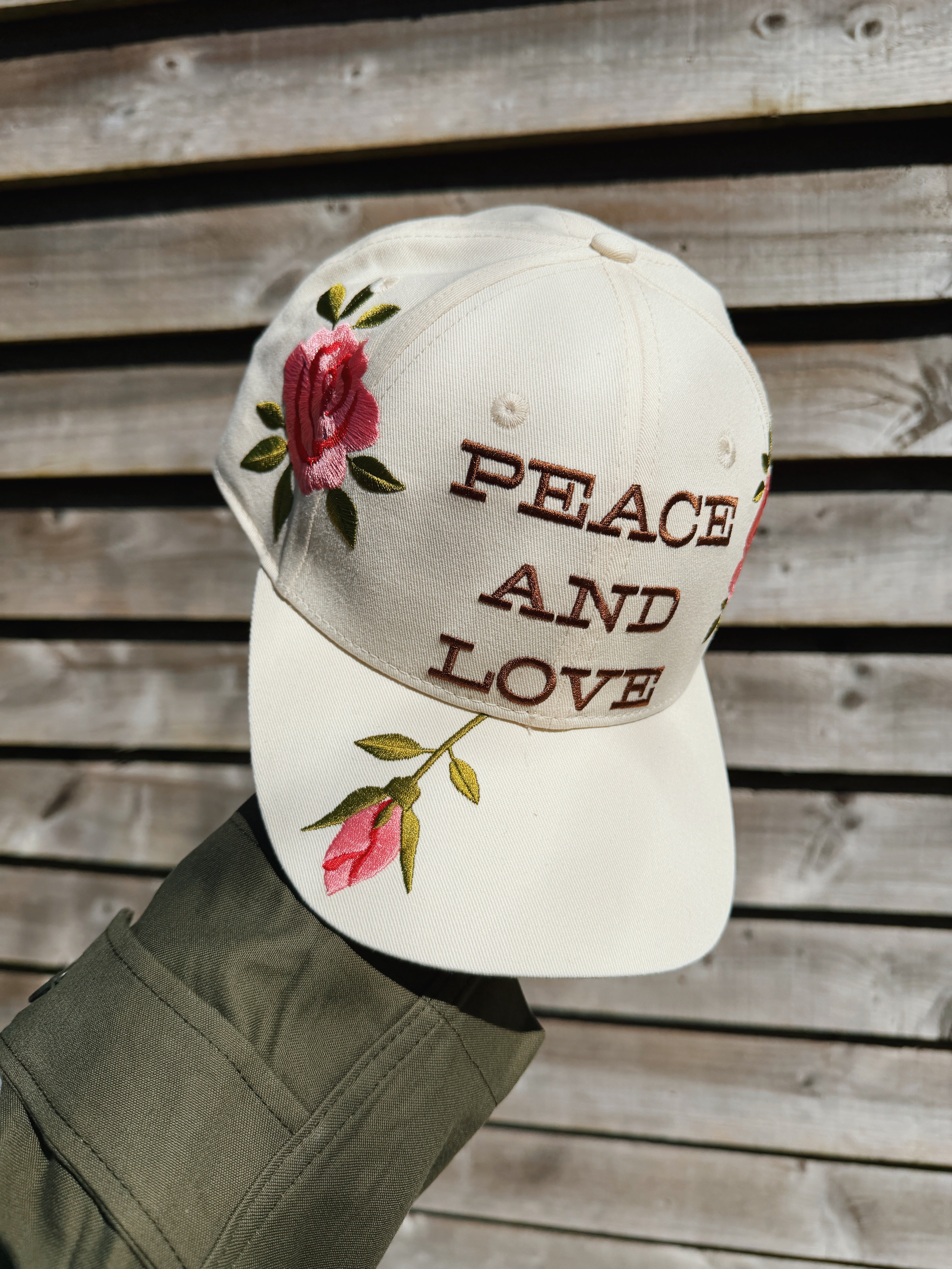 Cream baseball cap embroidered with "Peace and Love" lettering and pink rose floral details.
