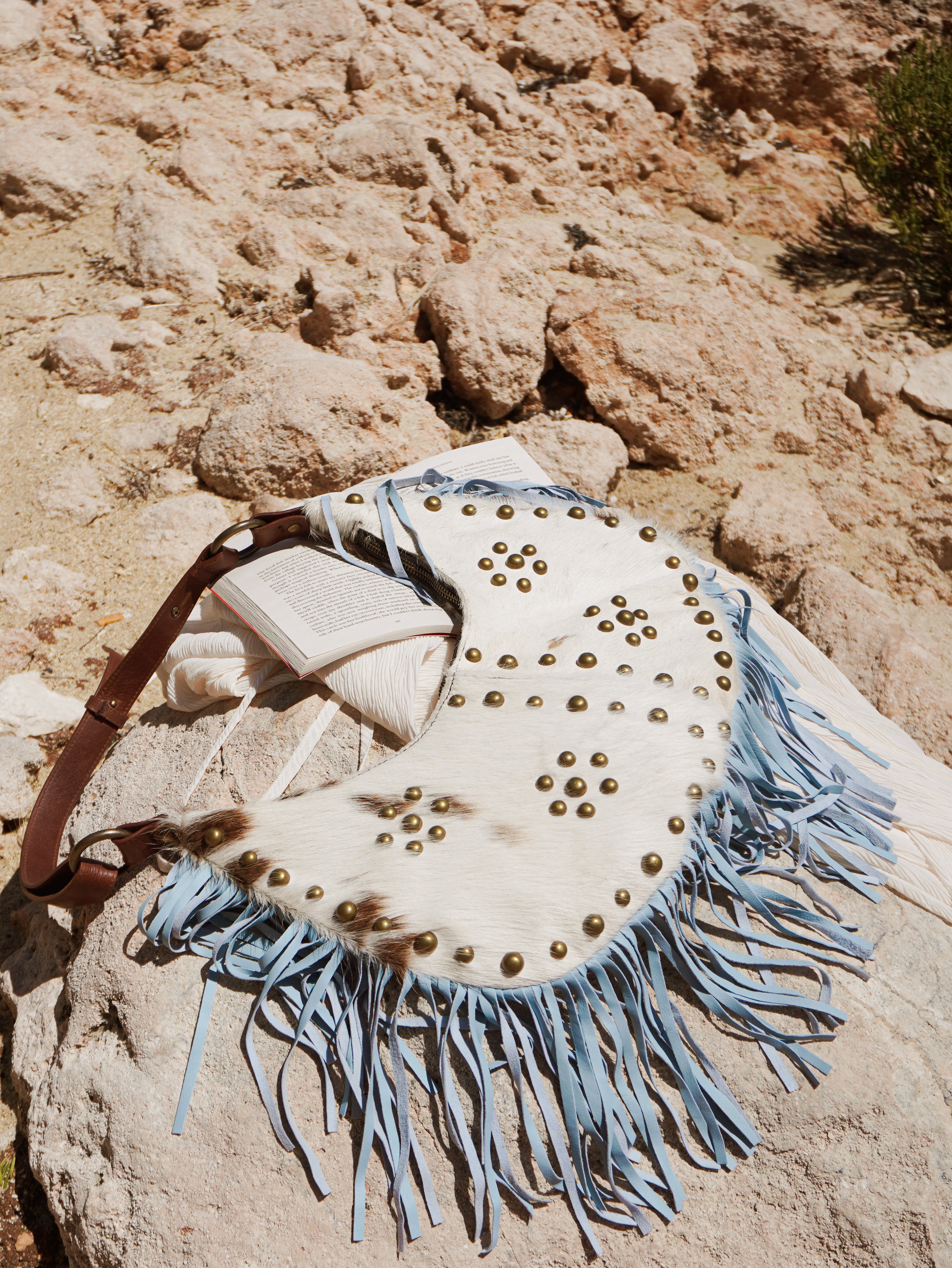 Cream and brown cowhide leather shoulder bag with light blue suede fringe and brass stud detailing resting on natural rocks.