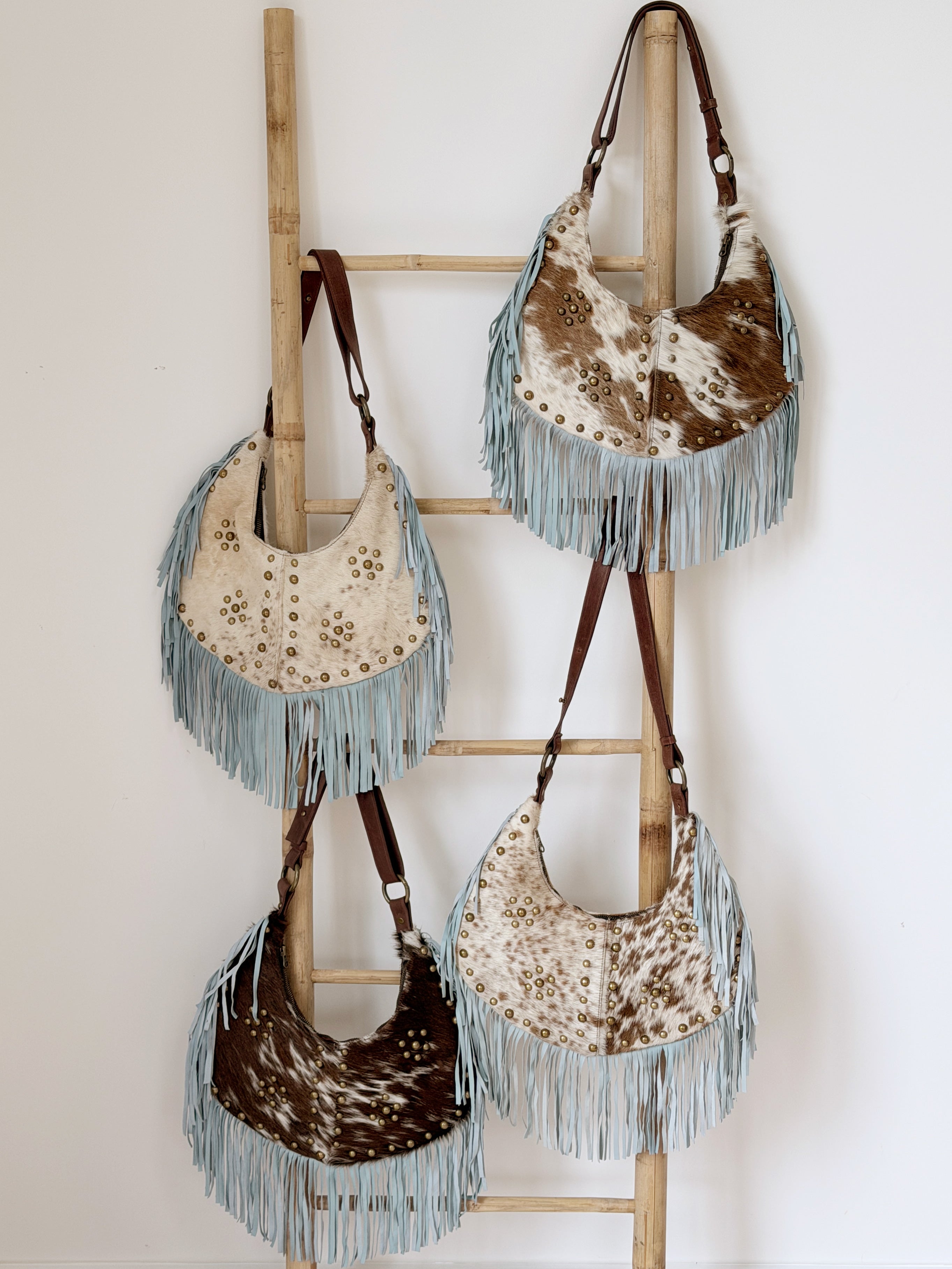 Collection of cowhide leather shoulder bags with light blue suede fringe, brass stud detailing, and brown leather straps.