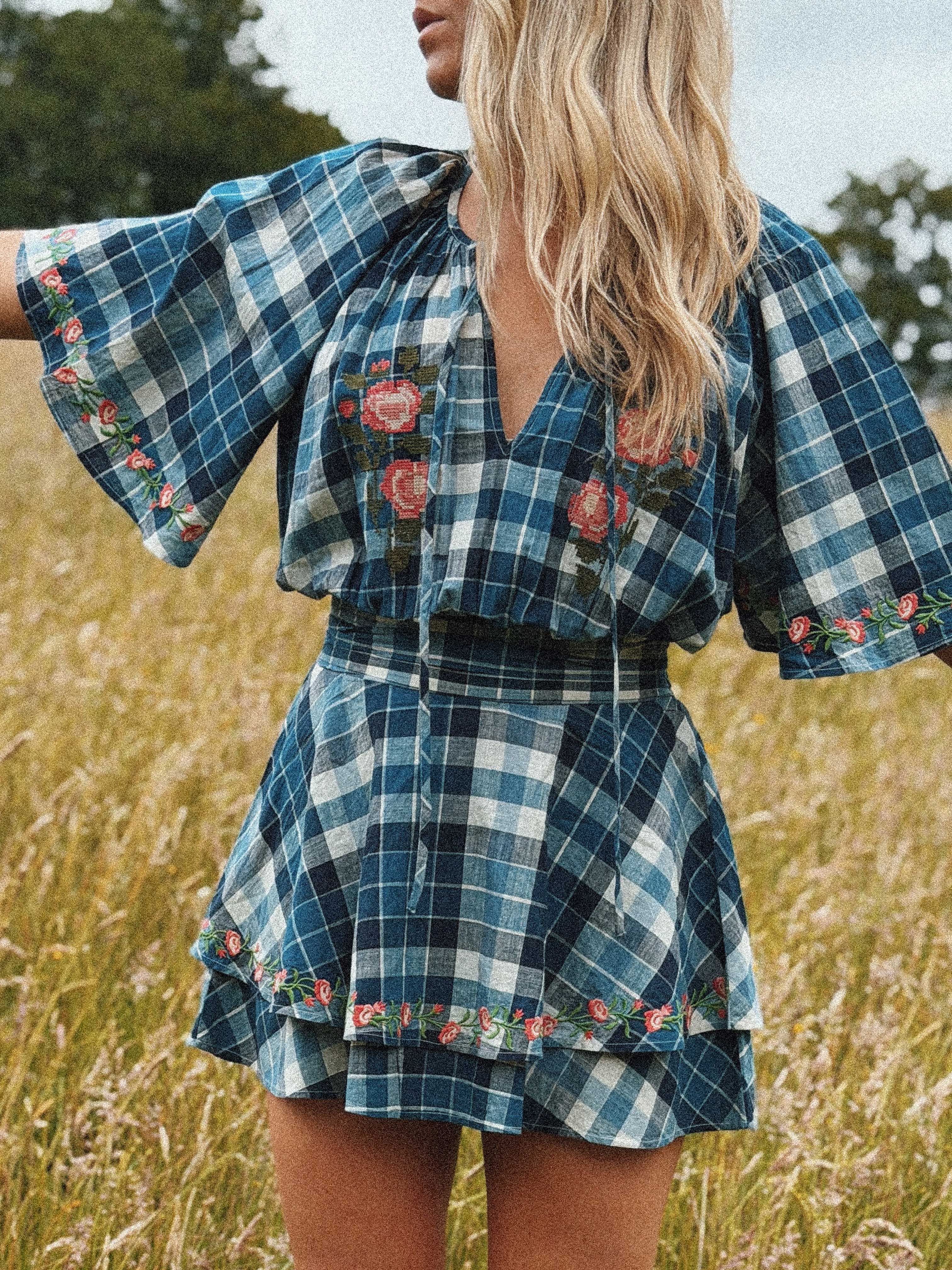 Person wearing a blue and white plaid mini dress with floral embroidery on the bodice, sleeves, and hem, featuring a V-neckline, flutter sleeves, and a tiered skirt.