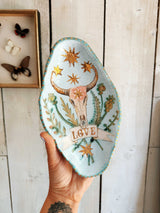 A hand holds a light blue ceramic dish decorated with a bull skull, gold stars, green leaves, and a banner reading 'LOVE'. The background is a white wooden wall with a framed butterfly display on the left.