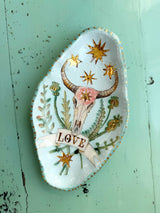 A decorative ceramic dish with a hand-painted western theme, featuring a bull skull, flowers, stars, and a banner reading 'LOVE', displayed on a distressed mint green wooden surface.