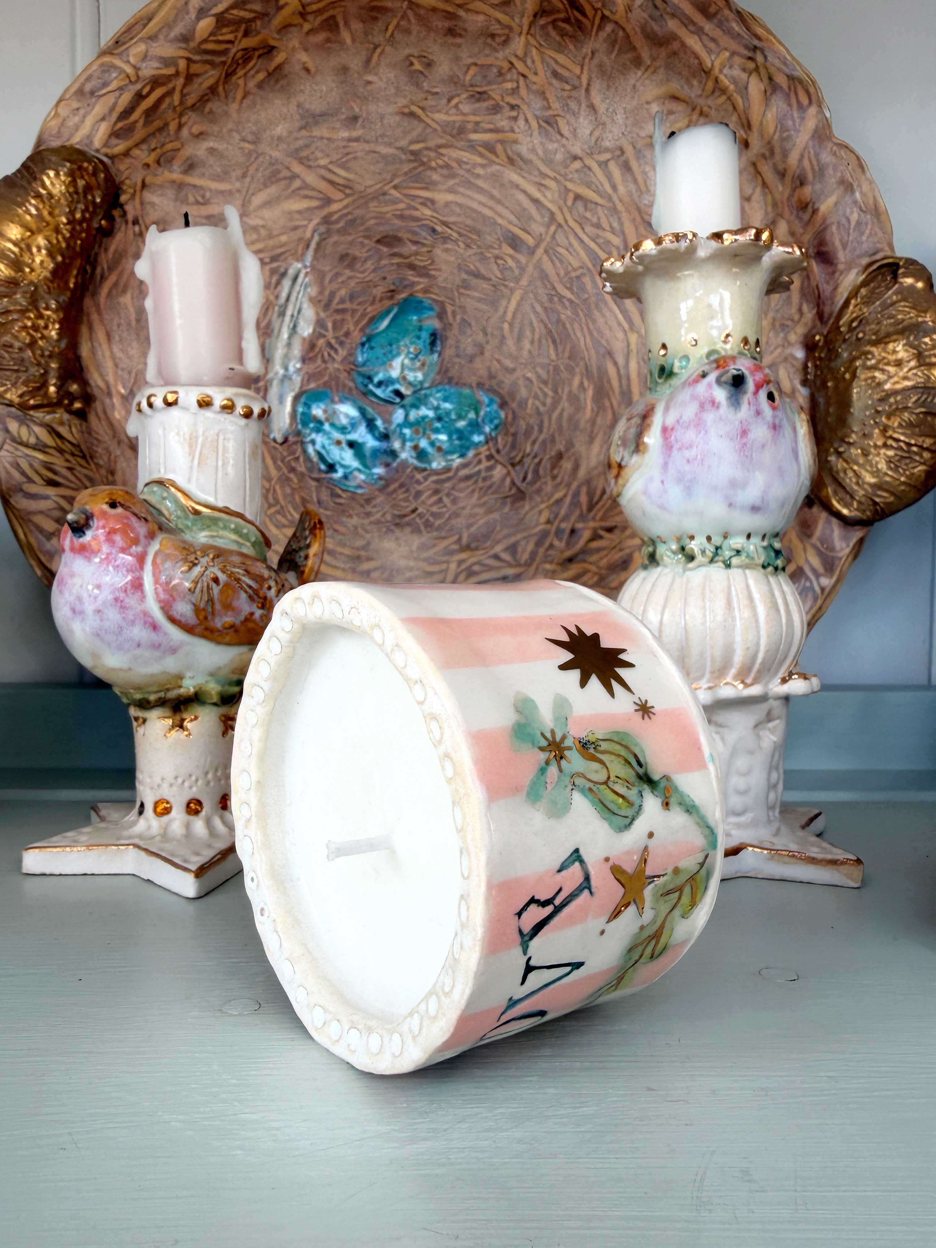 A decorative ceramic candle holder shaped like a bird with a pink and white striped body, green leaves, and a blue accent, shown with its candle removed and placed in front. Other matching bird and wing candle holders and a large woven tray are arranged behind it on a pale blue surface.