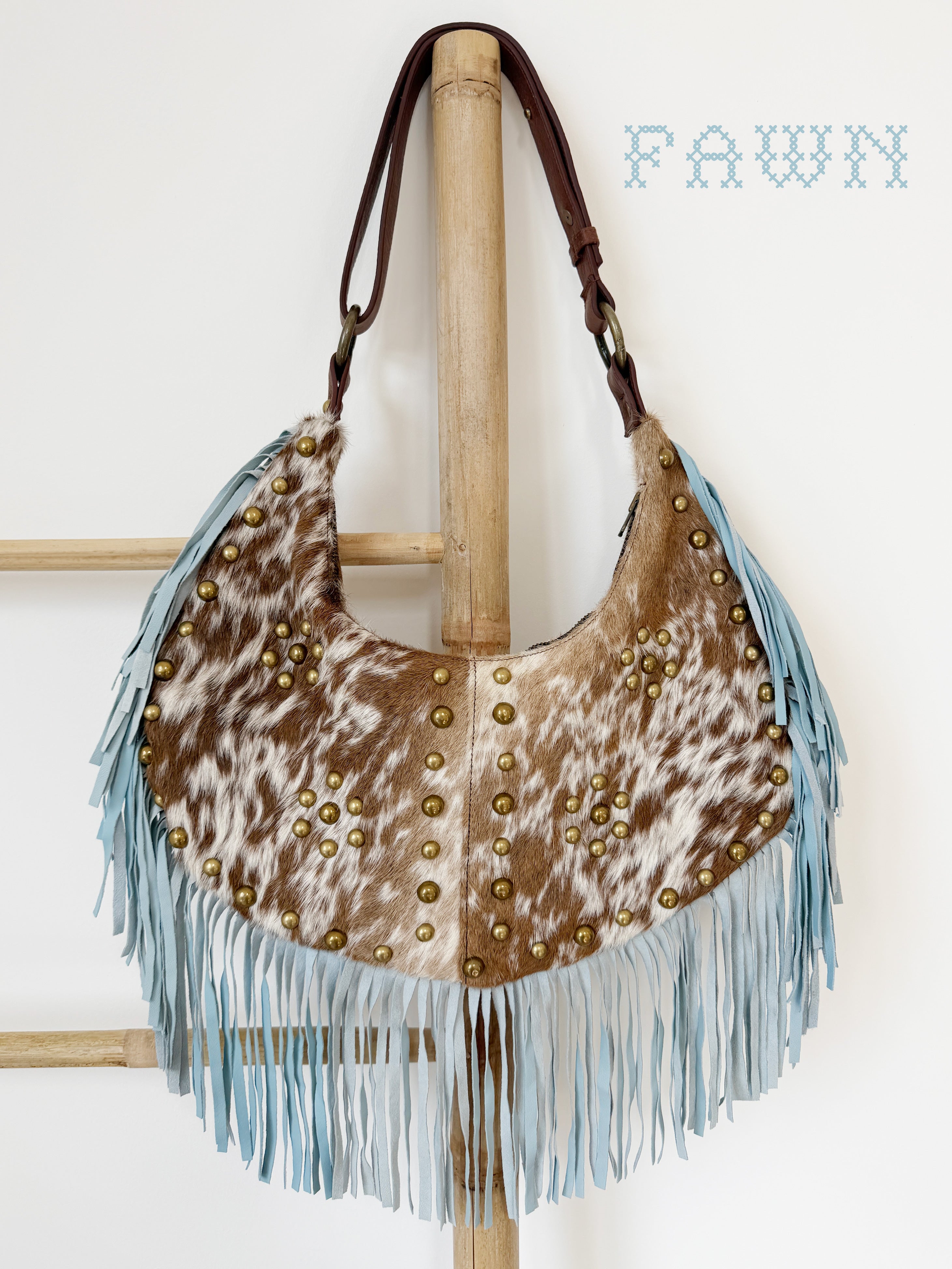 Fawn cowhide leather shoulder bag with light blue suede fringe, brass stud detailing, and a brown leather strap.