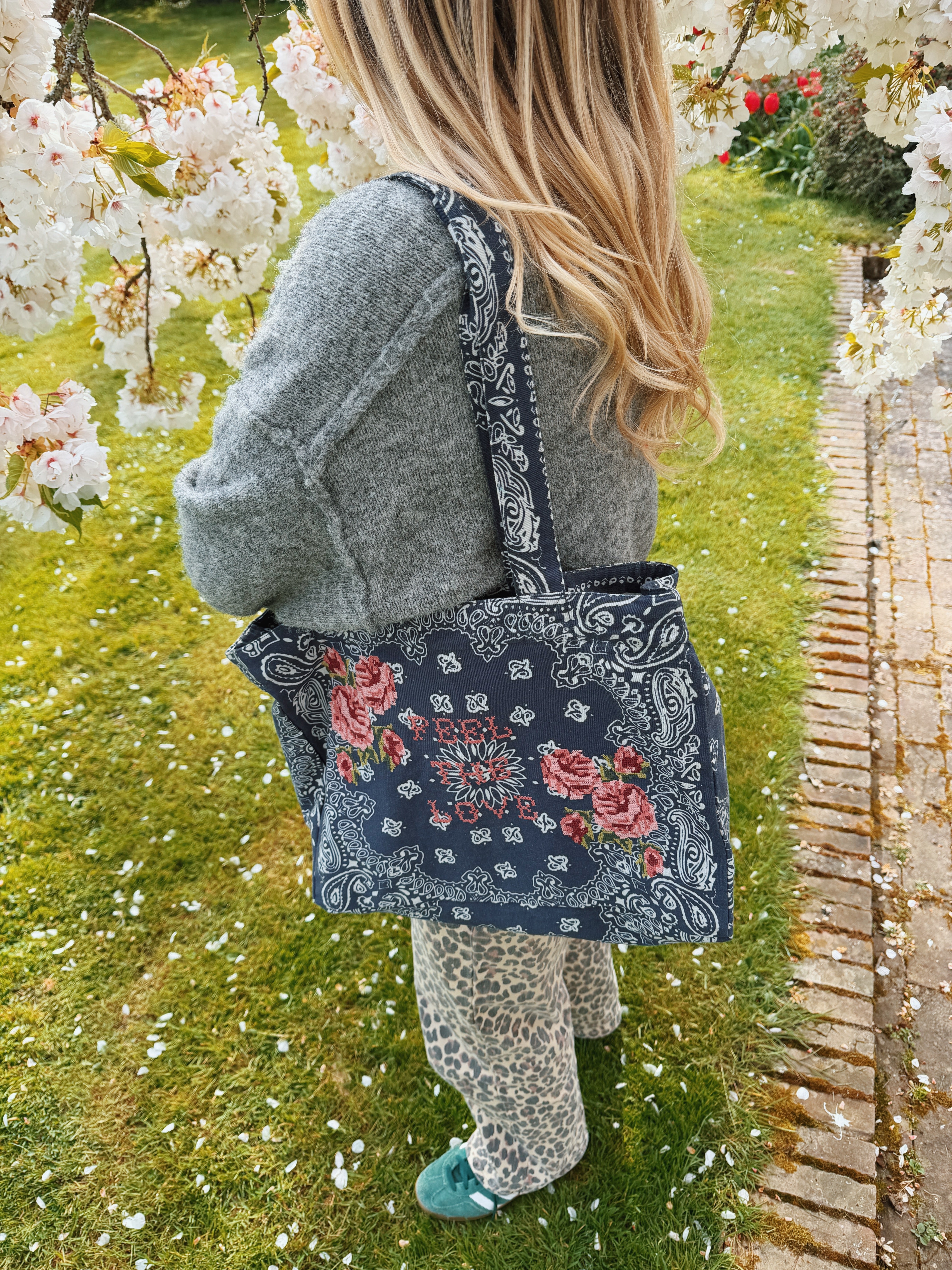 Person carrying a navy bandana print canvas tote bag embroidered with cross stitch roses and the words "Feel the Love."