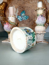 A decorative ceramic candle shown from the bottom, with a white wax interior and a painted exterior featuring the word 'LOVE' in blue letters, surrounded by flowers and leaves. It is styled on a tabletop alongside other vintage-style bird and candle holders.