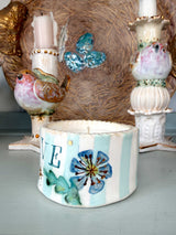 A handmade ceramic candle holder with a white and light blue striped body, decorated with a blue flower, green leaves, and the letter 'E'. It is styled on a light blue-gray surface with decorative bird candlesticks in the background.