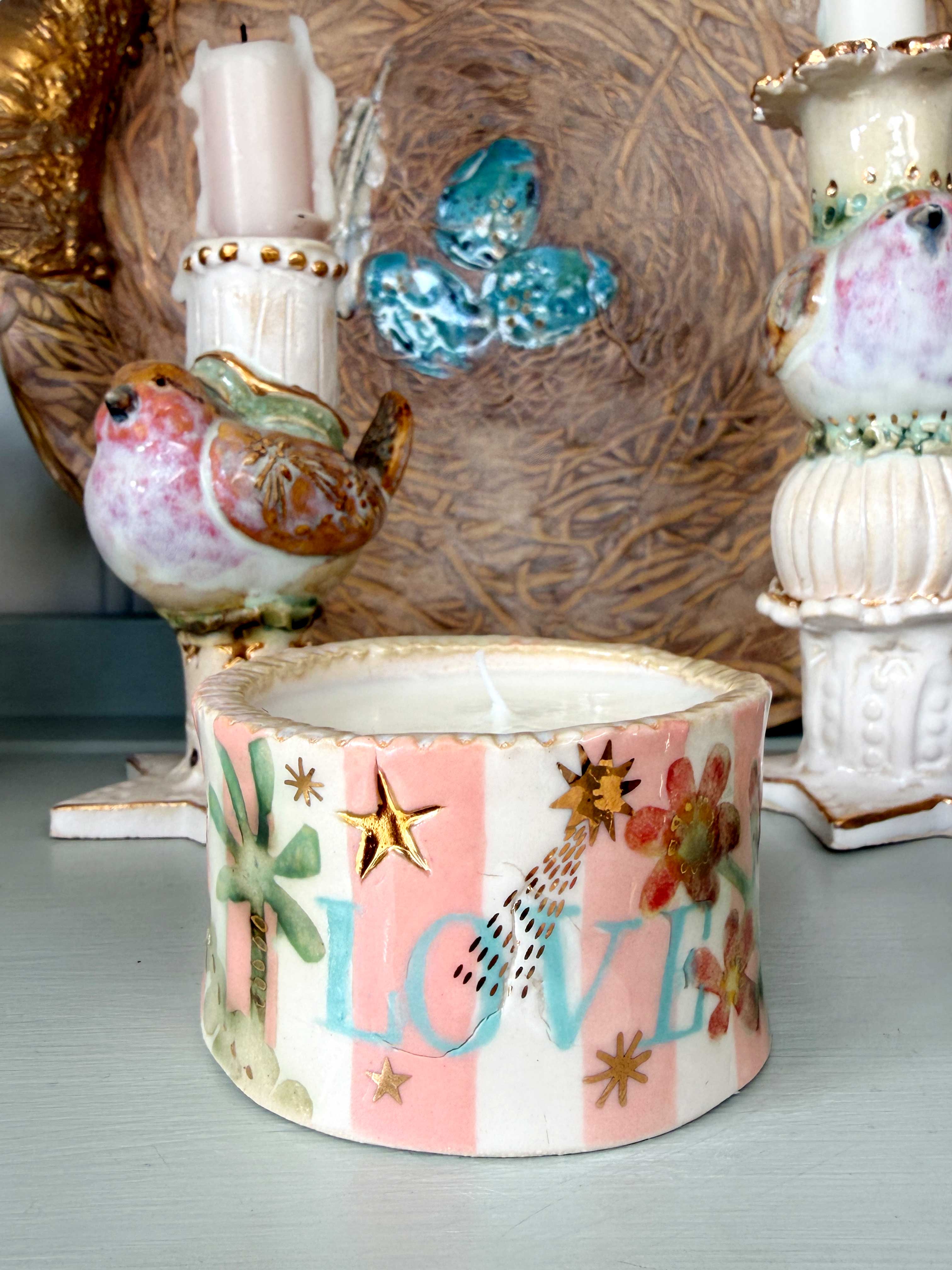 A small decorative ceramic candle holder with a pink and white striped pattern, gold stars, a bird design, and the word 'LOVE' in light blue, shown in a styled tabletop setting with other ceramic decor.