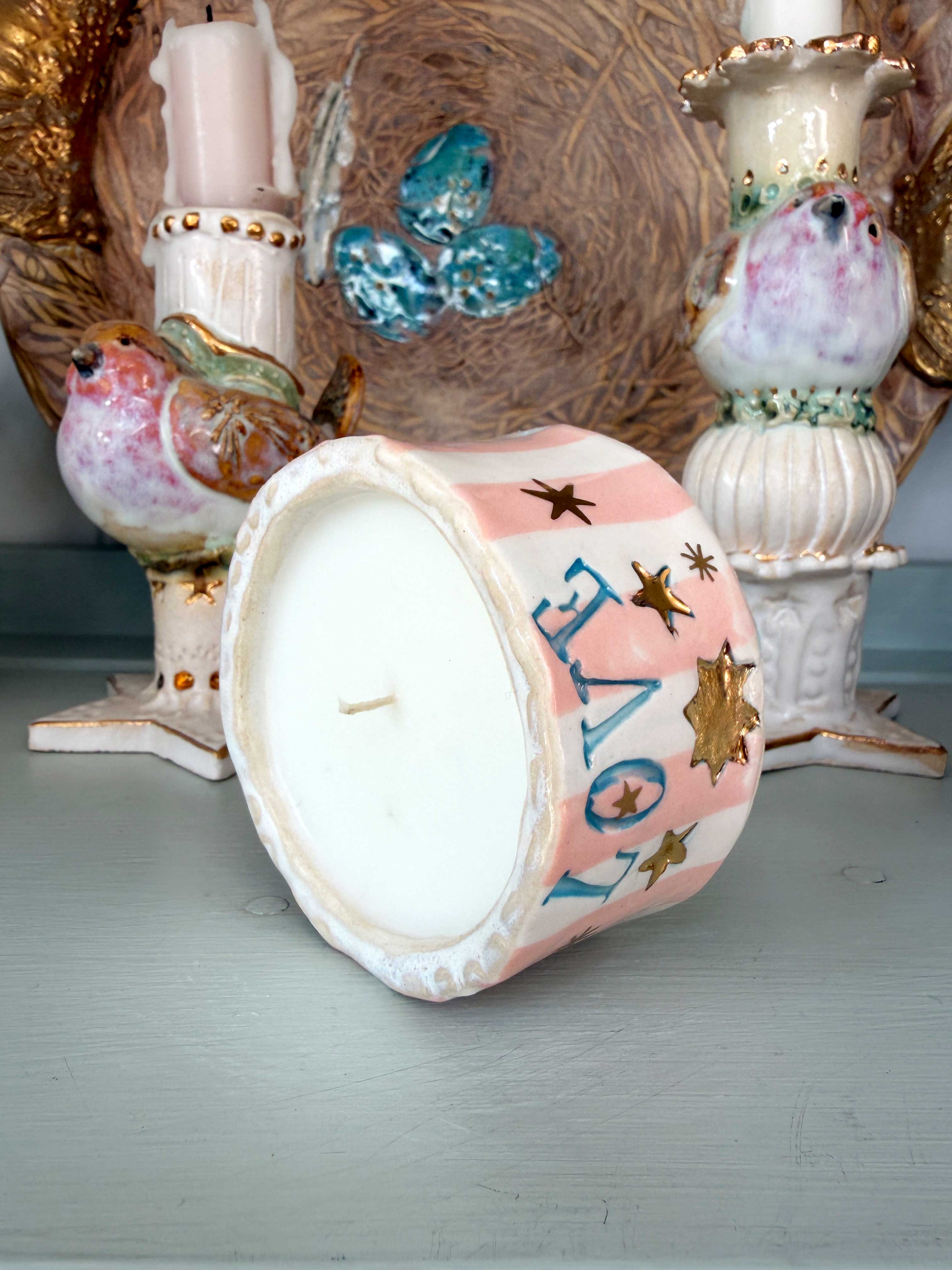 A hand-painted ceramic drum candle with a pink and white striped body, blue 'LOVE' lettering, and gold stars, shown lying on its side on a tabletop with decorative bird and candle figurines in the background.