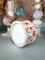 A hand-painted ceramic drum candle with a pink and white striped body, blue 'LOVE' lettering, and gold stars, shown lying on its side on a tabletop with decorative bird and candle figurines in the background.