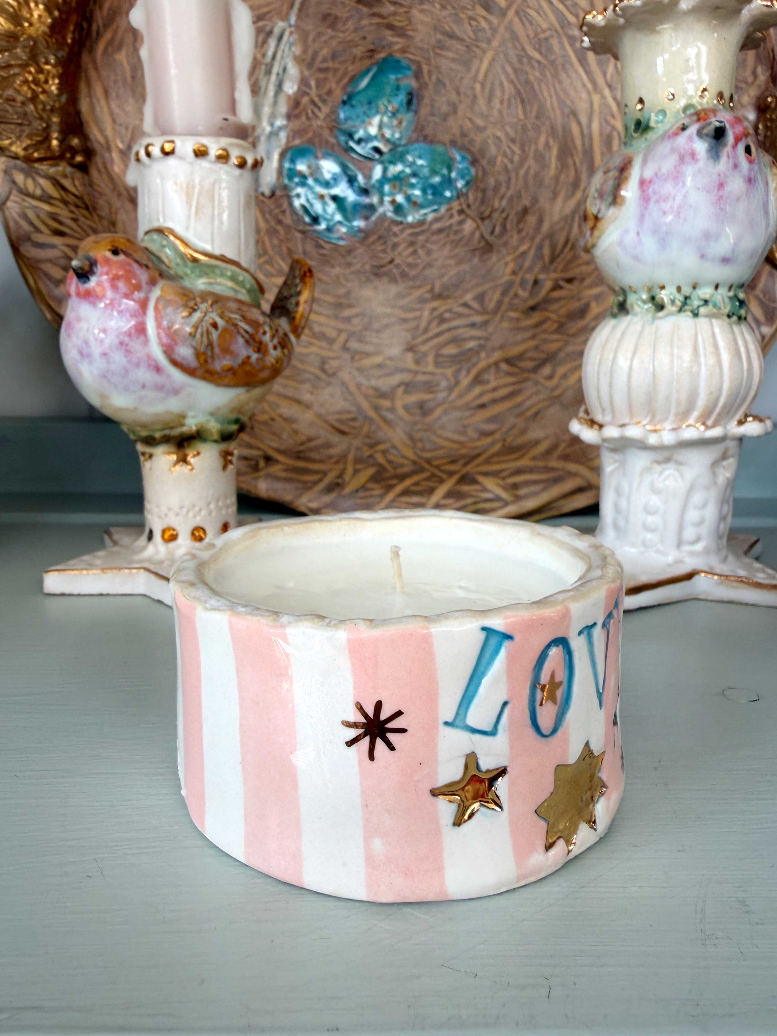 A handmade ceramic candle holder with pink and white vertical stripes, decorated with blue 'LOVE' lettering, gold and brown stars, and bird motifs, shown in a styled tabletop setting.