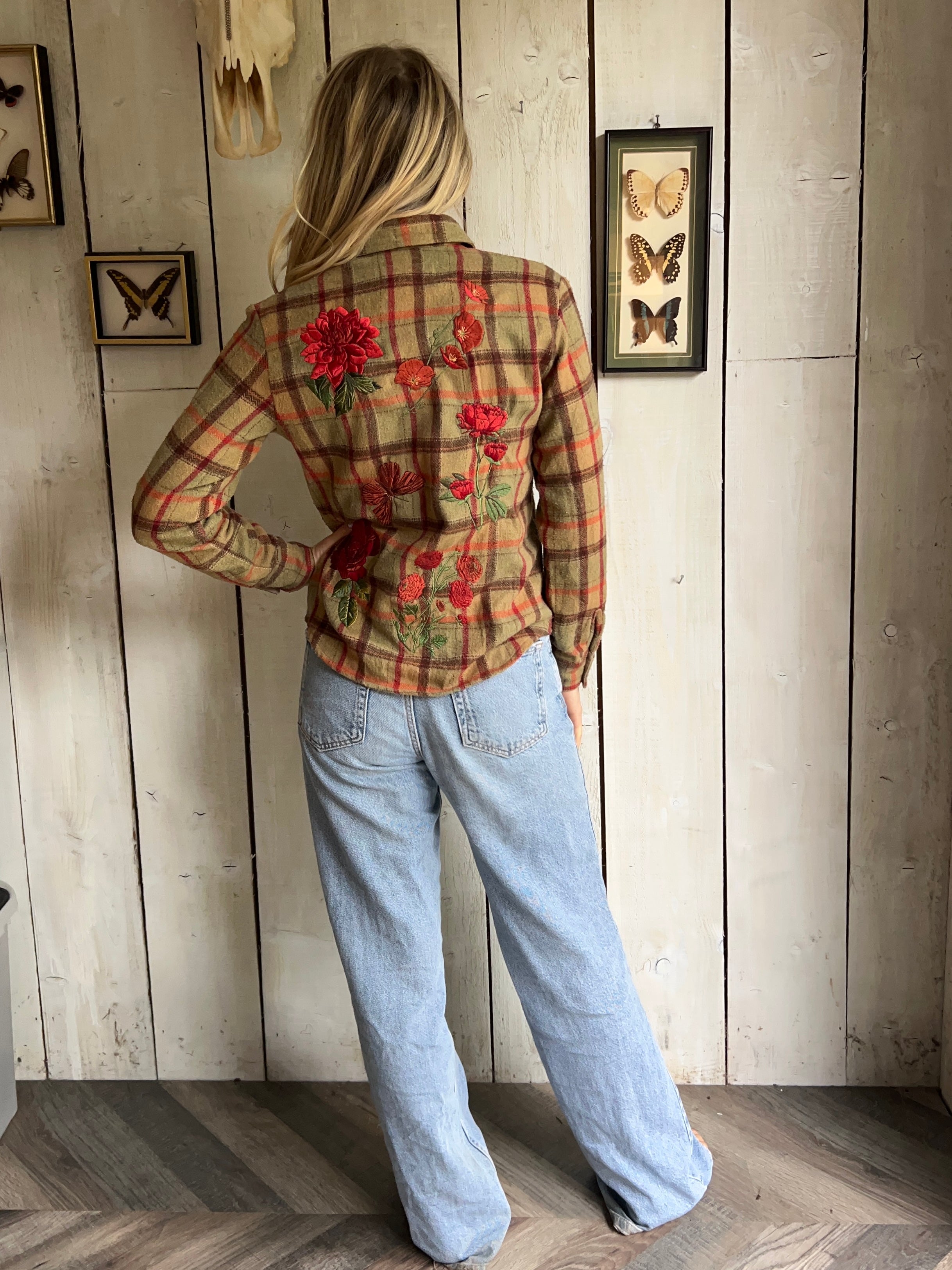 Person wearing a tan plaid shirt with red floral embroidery on the back, shown from behind.