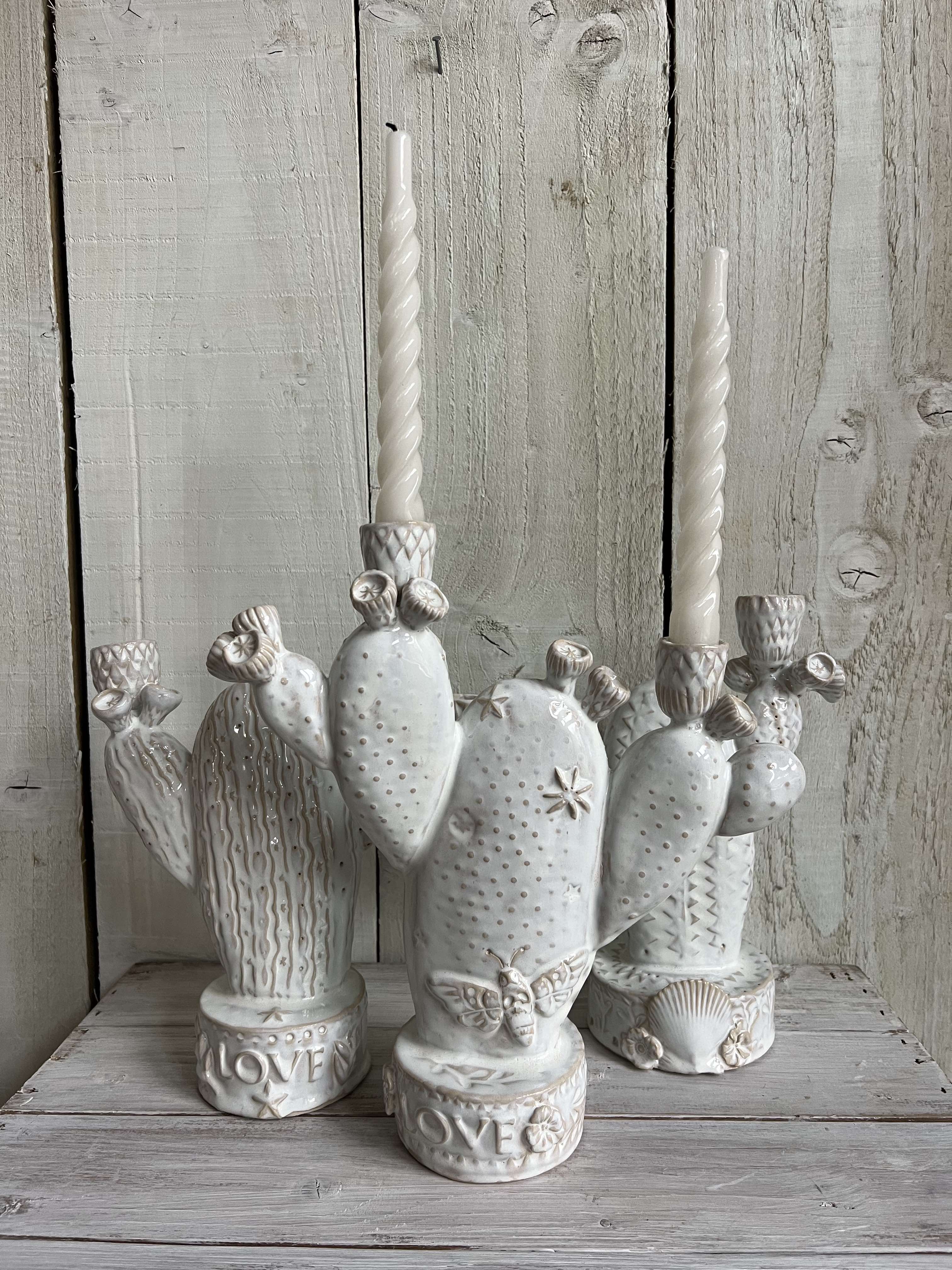 Collection of handmade white ceramic cactus candle holders displayed with taper candles on a rustic wooden shelf.
