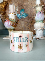 A handmade ceramic candle holder with the word 'SHINE' in blue, decorated with pink stripes, gold stars, and a gold crescent moon, styled on a tabletop with vintage bird candlesticks in the background.