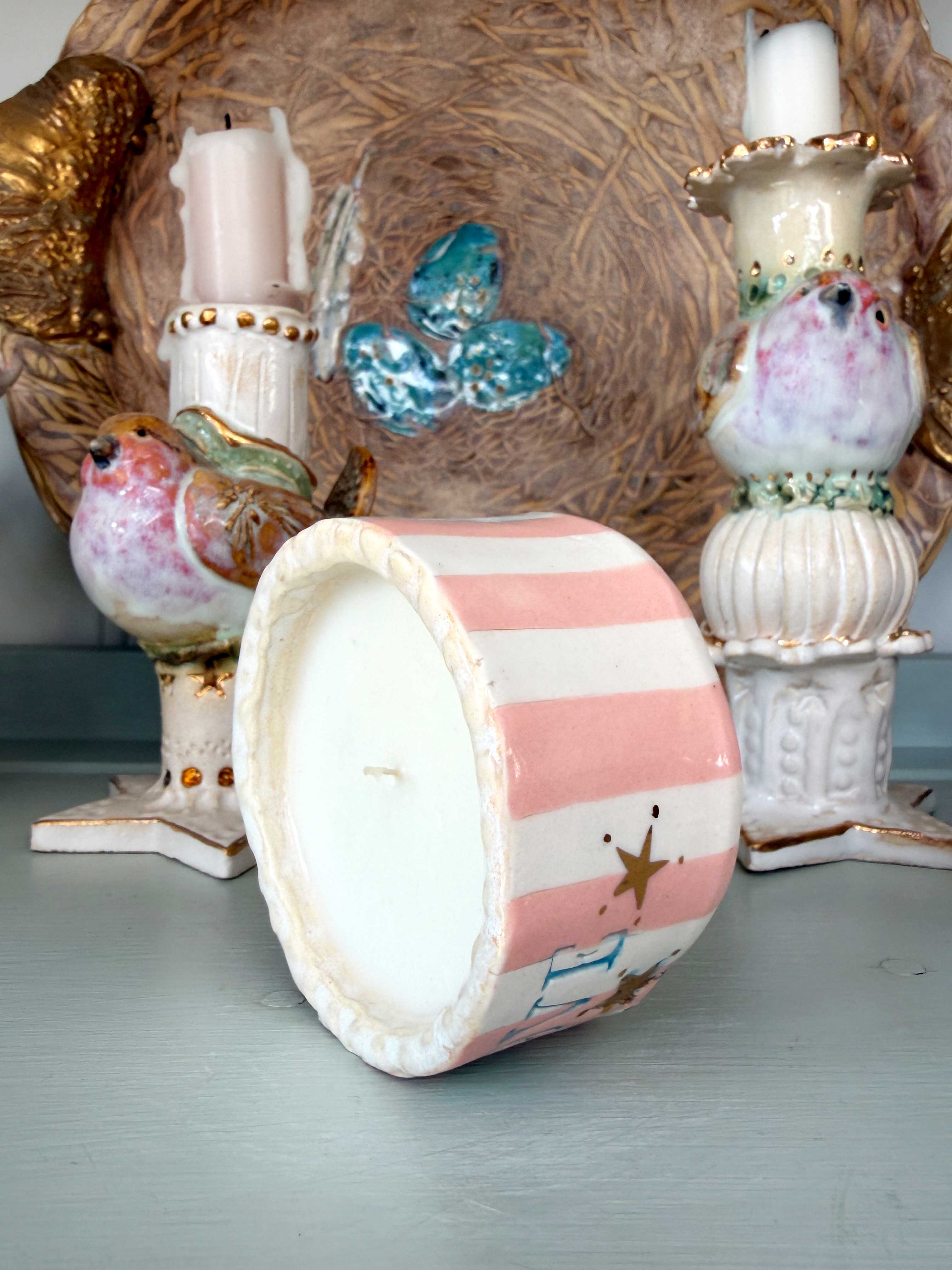 A decorative ceramic candle holder with pink and white horizontal stripes and gold star accents, shown lying on its side. The background features other vintage-style ceramic bird and birdhouse decorations on a painted gray surface.