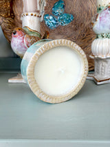 A ceramic candle jar with a cream-colored wax insert and a fluted rim, shown on a painted wooden surface with other decorative ceramic objects in the background.