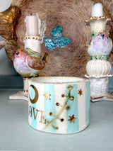A decorative ceramic candle holder with a bird and star design, set in front of other ornate ceramic bird candle holders on a light blue surface.