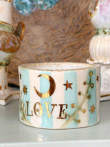 A cylindrical ceramic candle holder with a scalloped rim, featuring light blue and white vertical stripes, gold foil stars and a crescent moon, and the word 'LOVE' in gold. It is styled on a tabletop with other vintage ceramic items in a warm, softly blurred background.