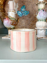 A handmade ceramic candle holder with vertical pink and cream stripes sits on a light gray tabletop, styled with vintage bird candlesticks and a decorative plate in the background.