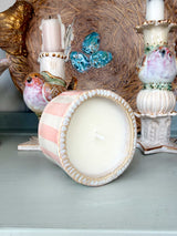 A decorative candle in a pastel pink and white striped ceramic pot with a scalloped rim, shown lying on its side on a tabletop. The candle is surrounded by other home decor items, including bird figurines, a blue butterfly ornament, and candlesticks.