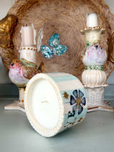A decorative ceramic candle holder with a bird design and floral patterns, shown with its lid placed in front of it. The candle holder is styled in a rustic holiday setting with other bird-themed candlesticks and a large woven basket in the background.