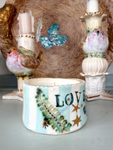 A handmade ceramic candle holder with a white and light blue striped base, decorated with a green pine tree, gold stars, and the word 'LOVE' in dark blue. It is styled on a tabletop with other decorative ceramic items in the background.