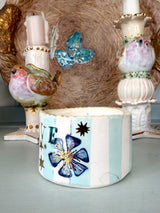 A handmade ceramic candle holder in the foreground, decorated with light blue and white vertical stripes, a blue flower with gold glitter accents, and a black star. In the background, two matching bird-shaped candlesticks and a rustic woven basket are visible.