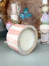 A pastel pink and white striped ceramic candle holder with a scalloped rim, shown tipped on its side to reveal a white candle inside. The background features other decorative ceramic items and a textured brown woven basket.