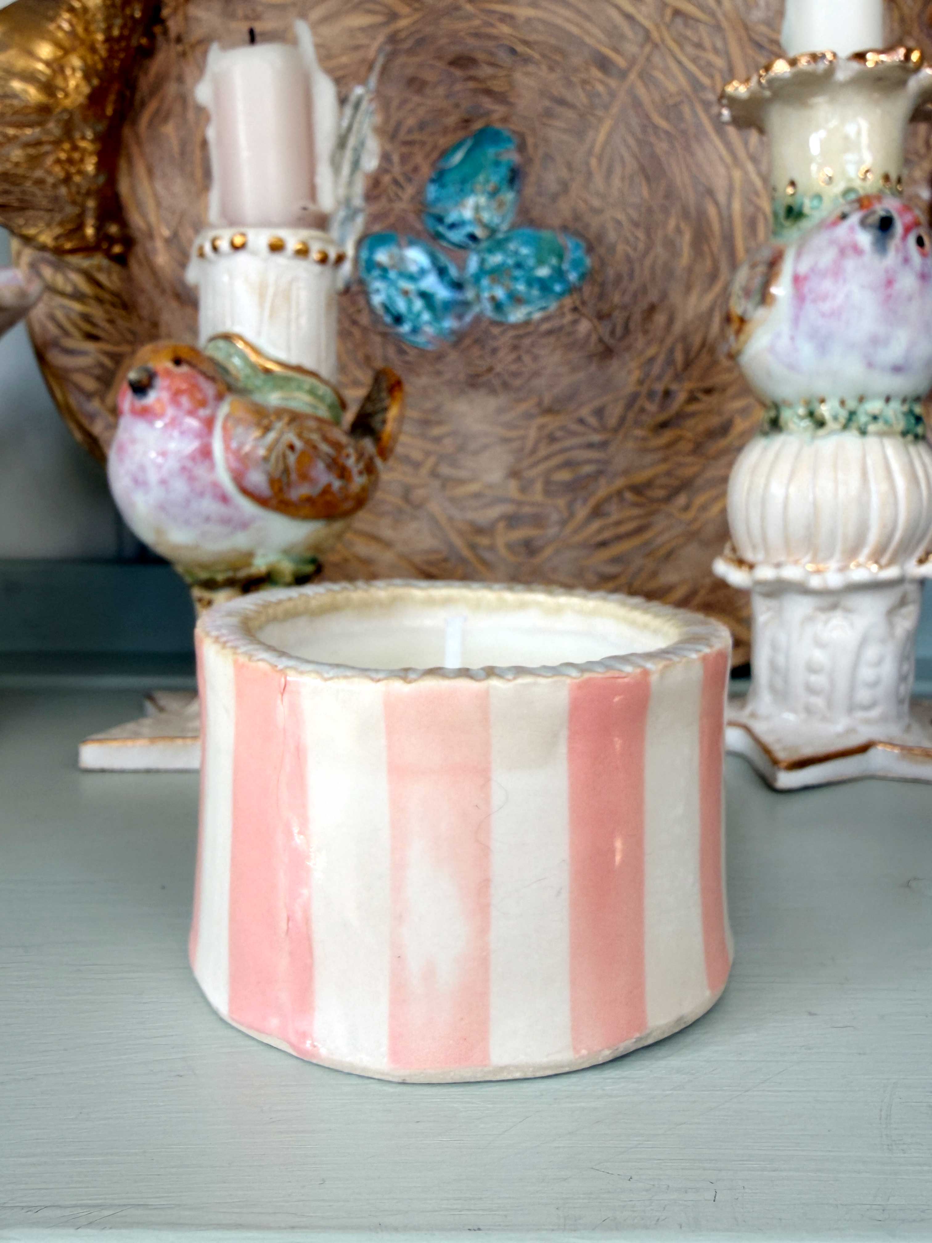 A decorative ceramic candle holder with pink and white vertical stripes, a pink flower motif, and gold stars, displayed on a shelf alongside other ornate ceramic candlesticks.