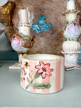 A decorative ceramic candle holder with pink and white vertical stripes, a pink flower motif, and gold stars, displayed on a shelf alongside other ornate ceramic candlesticks.