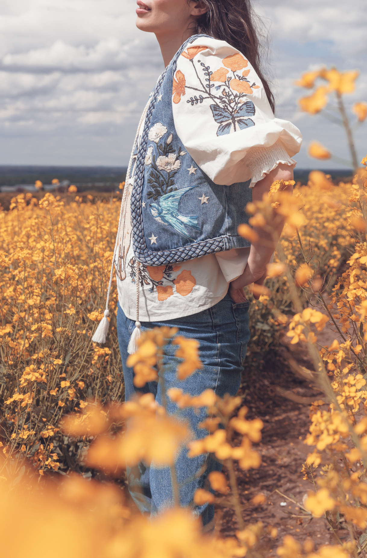 Person wearing a blue embroidered denim vest over a white embroidered blouse in a field of yellow wildflowers.