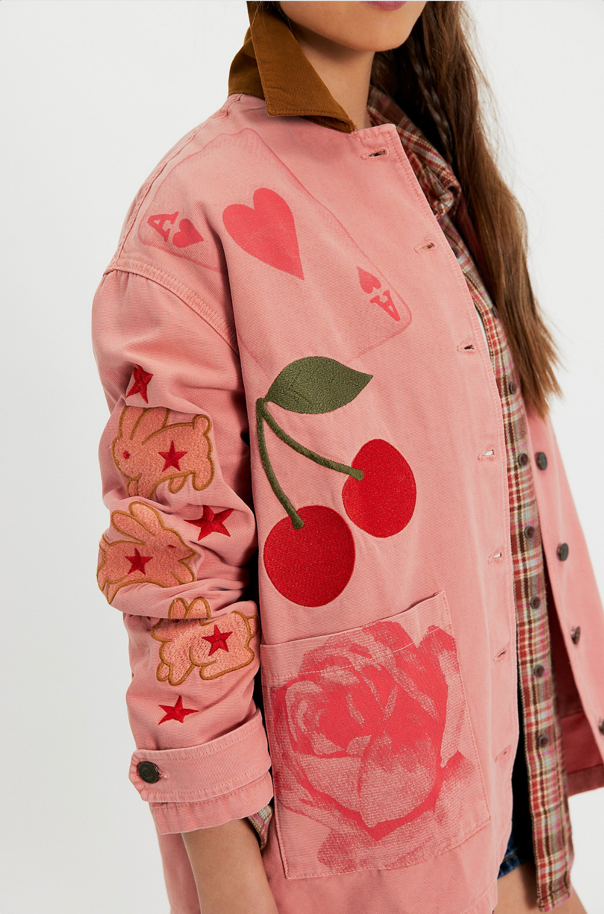 Woman wearing a pink hand-painted jacket featuring cherry, flower, heart, and bunny artwork over a plaid shirt.