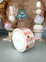 A decorative cylindrical candle with a white top and a pink-and-white striped side, decorated with small gold stars and blue script, shown lying on its side in front of ornate bird-shaped candle holders.
