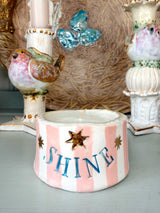 A handmade ceramic candle holder with pink and white vertical stripes, featuring the word 'SHINE' in blue lettering and gold star accents. The candle holder is styled on a tabletop with vintage bird candlesticks and a woven basket in the background.