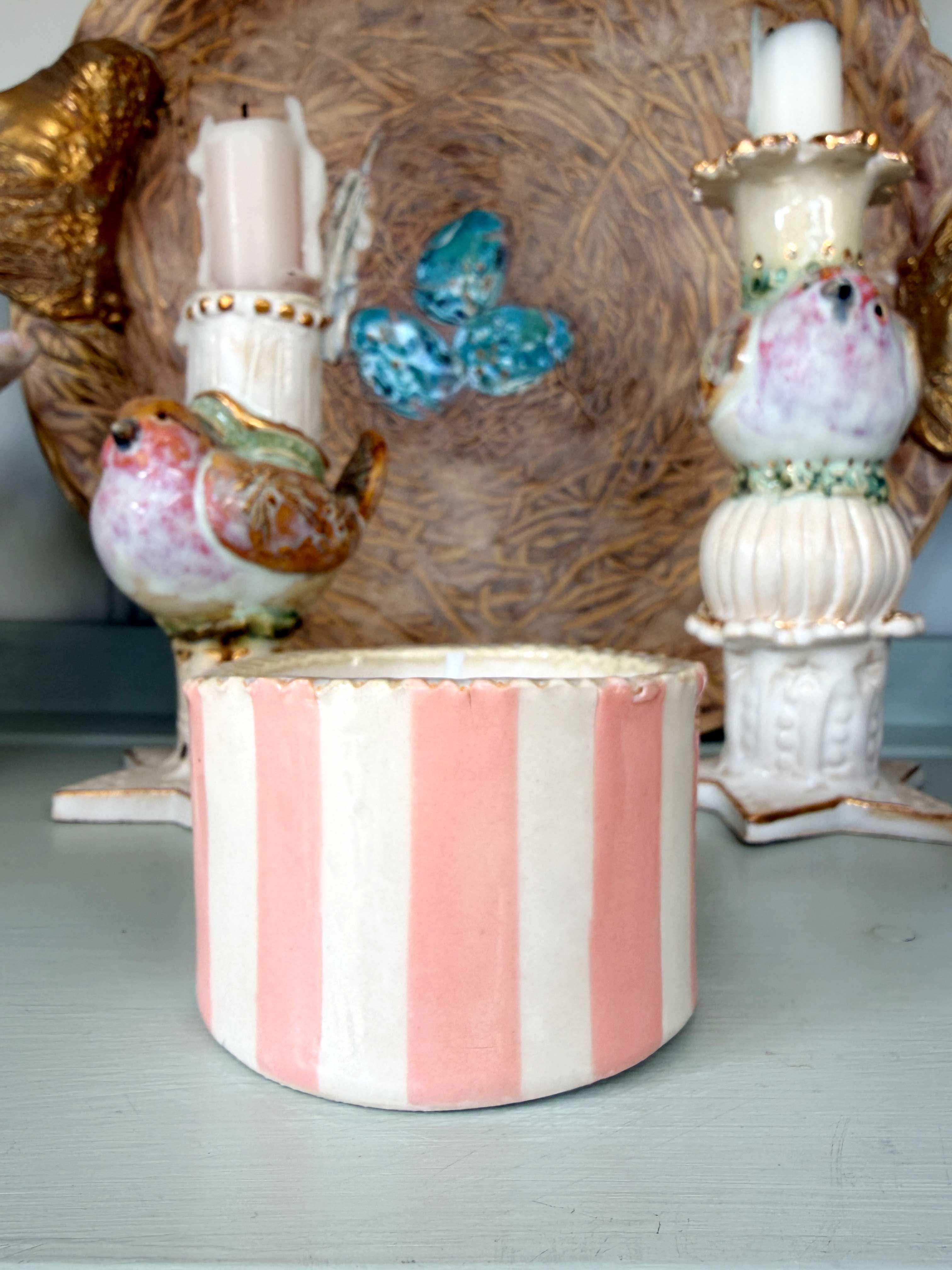 A cylindrical ceramic candle holder with vertical pink and white stripes, shown in front of a decorative vintage-style bird and butterfly centerpiece.