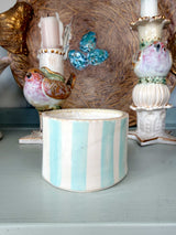 A cylindrical ceramic container with vertical light blue and white stripes, styled on a painted surface with decorative bird figurines and a textured background.