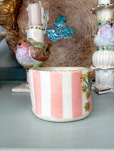 A small cylindrical ceramic container with vertical pink and white stripes and a gold floral accent, displayed on a tabletop with vintage-style decorative objects in the background.