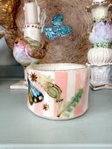 A handmade ceramic candle holder with pink and white vertical stripes, decorated with painted birds, butterflies, and leaves, displayed on a shelf among other decorative ceramic items.
