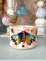 A small ceramic candle holder with pink and white stripes, decorated with a colorful painted moth and gold stars, displayed on a shelf among other vintage-style decorative items.