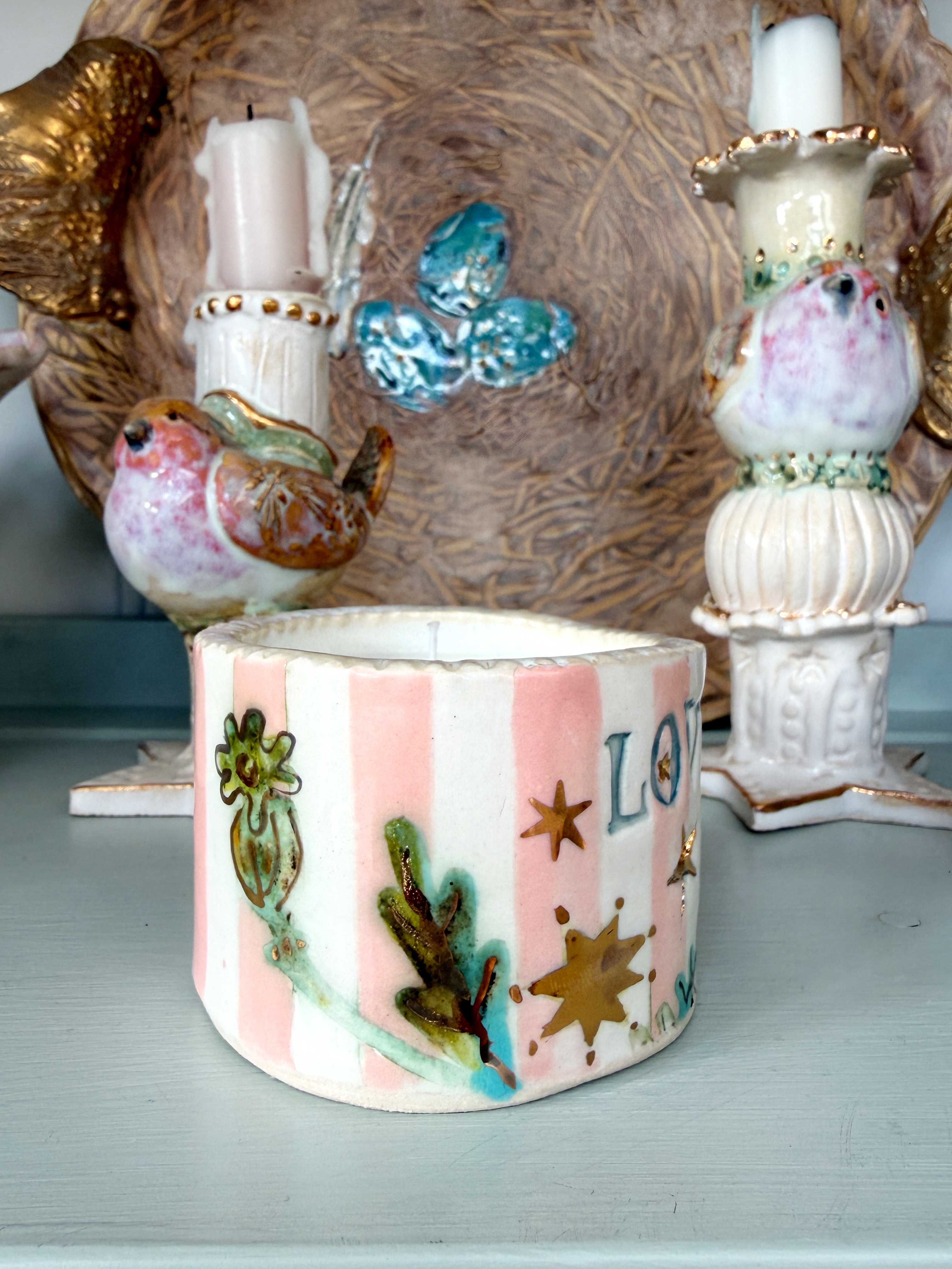 A handmade ceramic candle holder with pink and white vertical stripes, decorated with hand-painted green leaves, gold stars, and the letter 'LO'.
