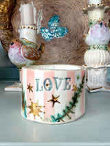 A handmade ceramic container with the word 'LOVE' in raised blue letters, decorated with pink and white stripes, gold stars, and green pine branches, displayed on a shelf with vintage bird candleholders.