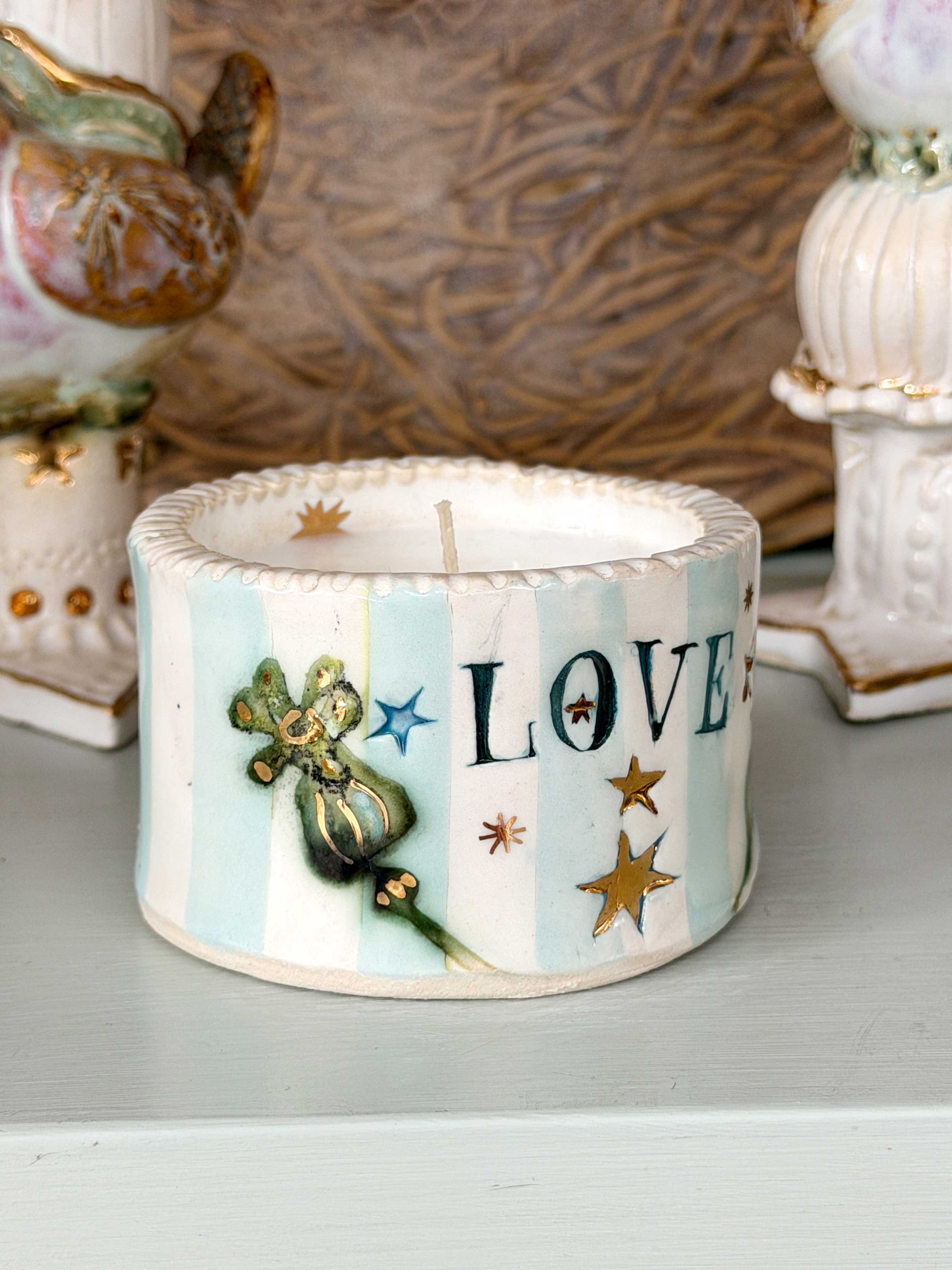 A small decorative ceramic candle holder with a white and light blue striped pattern, featuring the word 'LOVE' in dark blue letters, gold stars, and a green fairy illustration. The candle is shown on a tabletop with other vintage-style ornaments blurred in the background.