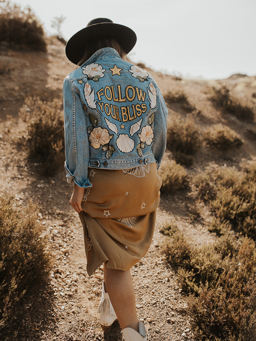 Light blue denim jacket with "Follow Your Bliss" embroidered back design, floral embroidery, and celestial motifs, shown from the back.
