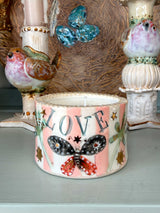 A handmade ceramic candle holder with a pink and white background, decorated with a black and red butterfly, gold stars, and the word 'LOVE' in blue letters. The candle is shown in a styled setting with other vintage bird figurines and decorative items.
