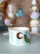 A handmade ceramic candle holder with vertical light blue and white stripes, decorated with a brown crescent moon, blue lettering, and green leaf accents, displayed on a light blue surface with decorative bird candlesticks in the background.