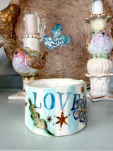 A handmade ceramic candle holder with the word 'LOVE' painted in blue, decorated with colorful bird and butterfly motifs, set against a rustic background of other decorative home items.