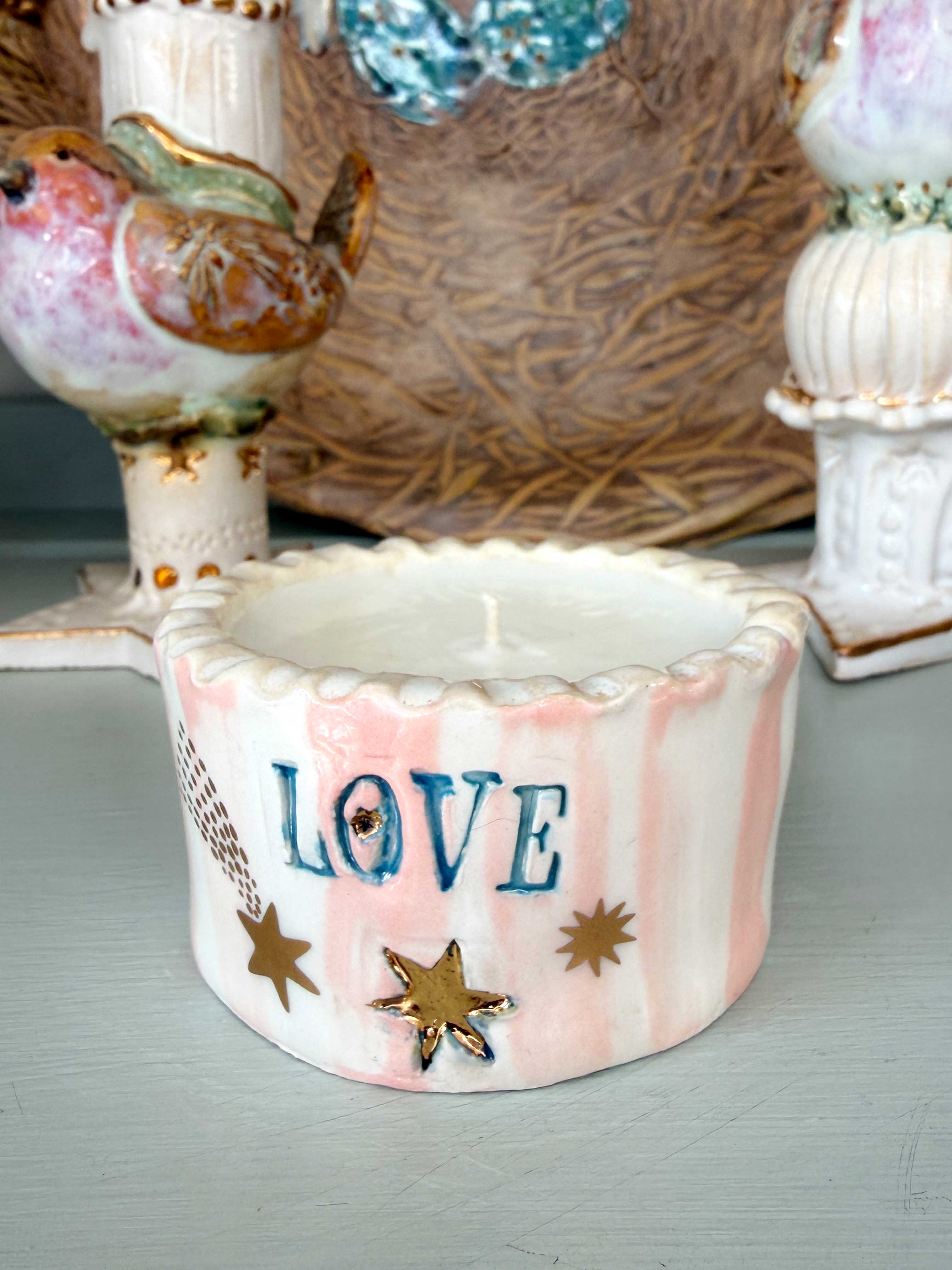 A small white ceramic candle holder with pink vertical stripes, blue 'LOVE' lettering, and gold star accents, displayed on a tabletop with decorative bird figurines in the background.