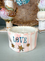 A small white ceramic candle holder with pink vertical stripes, blue 'LOVE' lettering, and gold star accents, displayed on a tabletop with decorative bird figurines in the background.