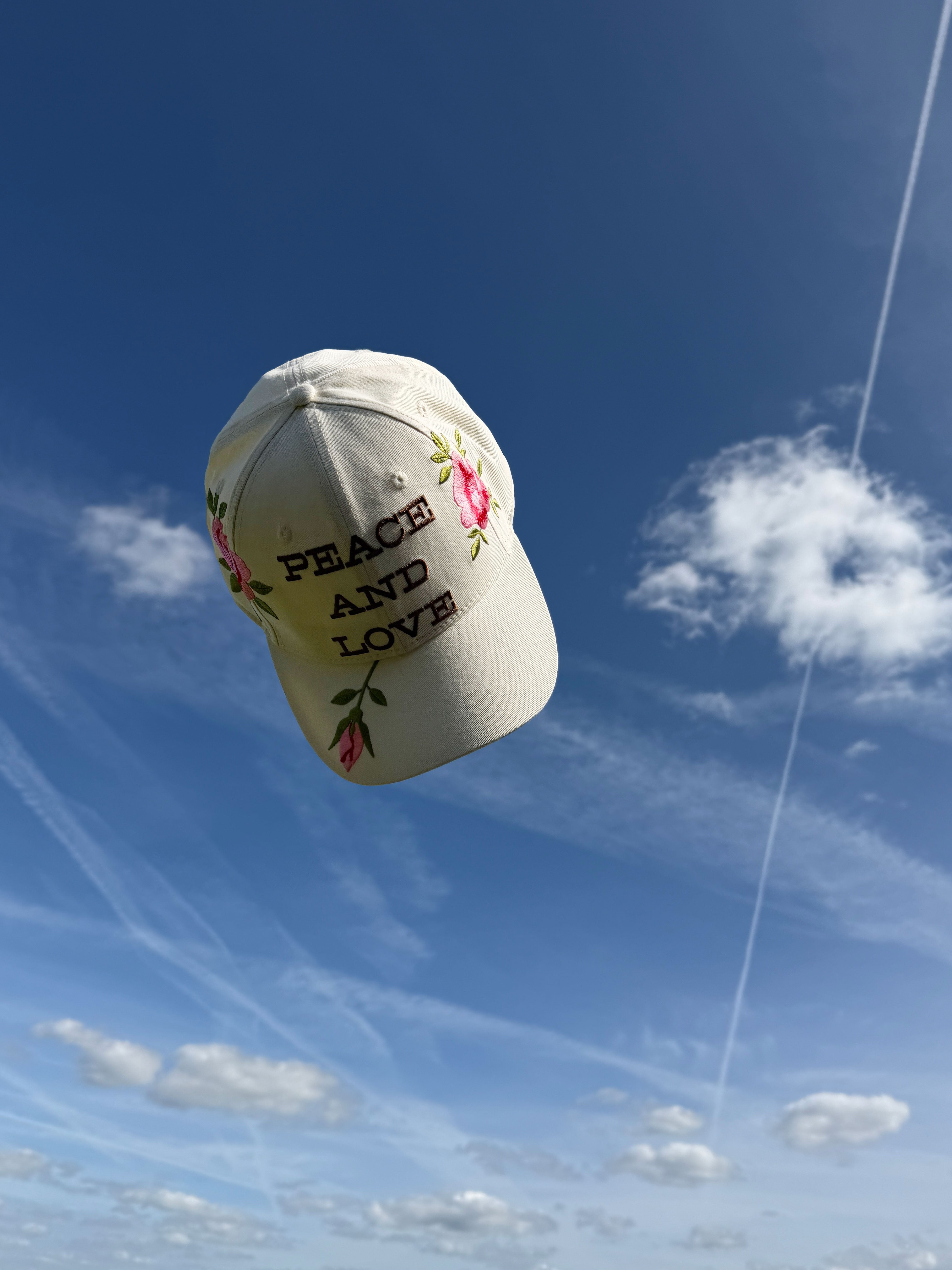Cream baseball cap embroidered with "Peace and Love" and pink roses against a blue sky.