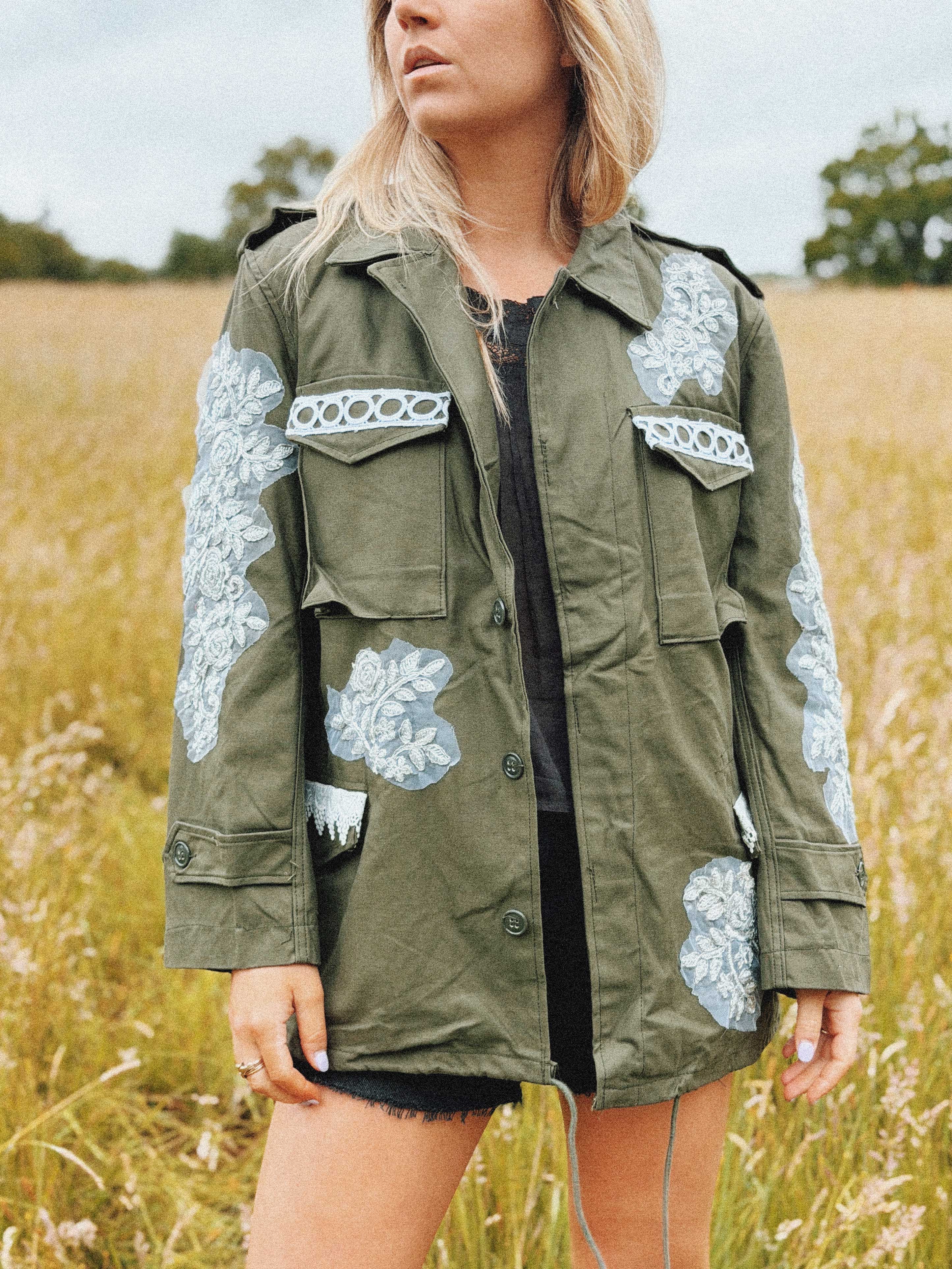 Olive green military jacket with white floral lace appliqué and decorative trim, front view.